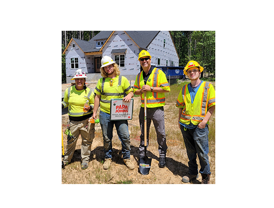 4 people wearing hard hats and yellow safety vests in front of a home construction site with shovel and pizza box